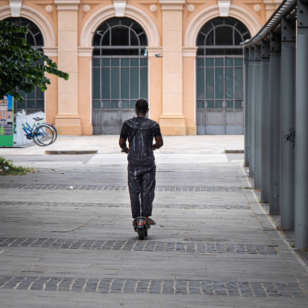 Μαύρος άνδρας ντυμένος σε γκρι Διασχίζοντας μια δημόσια πλατεία για το ηλεκτρικό Mono σκούτερ. - Φωτογραφία, εικόνα
