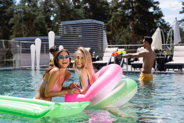 aufgeregte Frauen lachen in die Kamera, während sie sich im Schwimmbad neben verschwommenen multiethnischen Freunden ausruhen - Foto, Bild