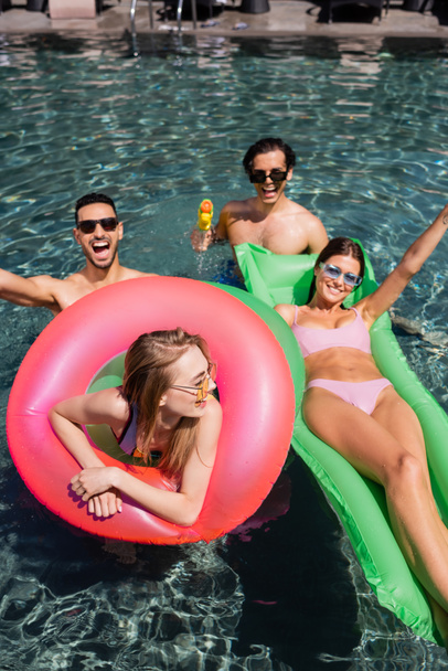 happy women on inflatable mattress and swim ring resting in pool near multiethnic men - 写真・画像