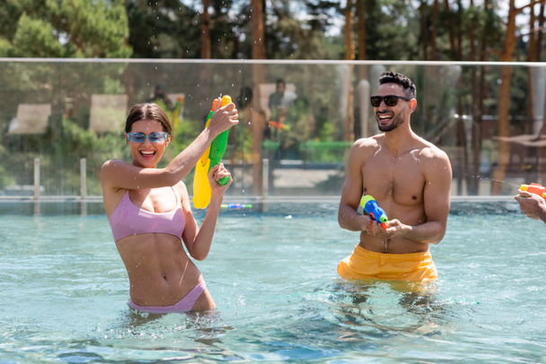 cheerful multicultural friends playing with water guns in swimming pool - Fotó, kép