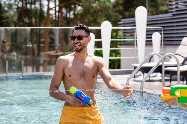cheerful arabian man holding water pistol in swimming pool - Photo, Image