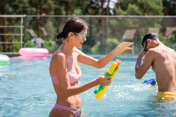 młoda kobieta z pistoletem wodnym gestykulująca blisko arabskiego przyjaciela w basenie - Zdjęcie, obraz