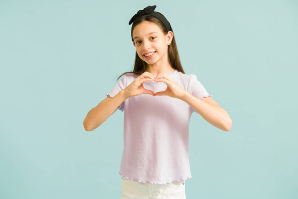 Cute happy little girl smiling while making a heart gesture with her hands. Portrait of a caucasian preteen girl sending love against a green background - Valokuva, kuva
