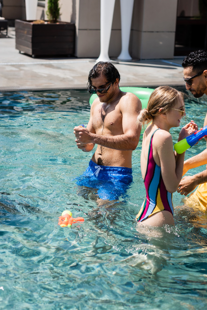 багатонаціональні чоловіки з молодою жінкою грають з водяними гарматами в басейні
 - Фото, зображення