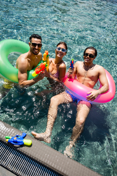 vista de alto ângulo de felizes amigos inter-raciais com anéis de natação e armas de água na piscina - Foto, Imagem