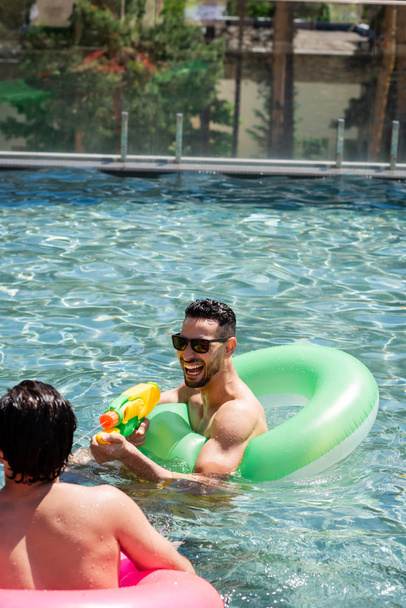 radosny arabski mężczyzna w okularach przeciwsłonecznych bawiący się pistoletem na wodę w pobliżu przyjaciela w basenie - Zdjęcie, obraz