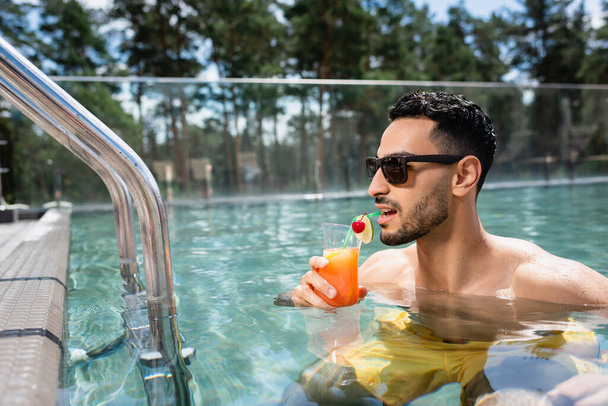 young muslim man in sunglasses drinking cocktail in swimming pool - Photo, Image