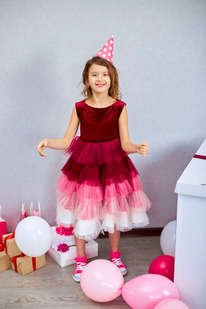 Söpö, iloinen pieni tyttö vaaleanpunainen mekko ja hattu pelata ilmapalloja kotona syntymäpäiväjuhlat serpentiinit, Hyvää syntymäpäivää. Juhlitaan. - Valokuva, kuva