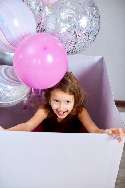 Cute, joyful little girl in pink dress looks out of the big present gift box with balloons at home birthday party streamers, Happy birthday. Celebrating - Fotó, kép