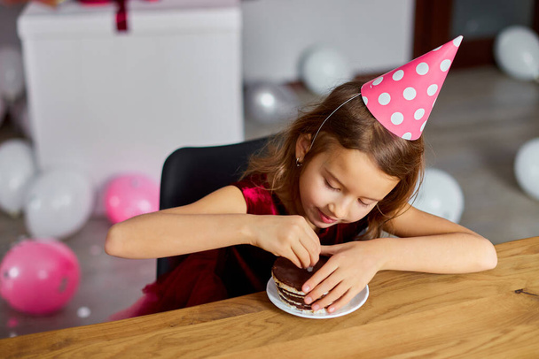 Mała szczęśliwa dziewczyna nosi kapelusz urodzinowy smaczny świeca z tortu urodzinowego, zrobić twarze, koncepcja uroczystości party. - Zdjęcie, obraz
