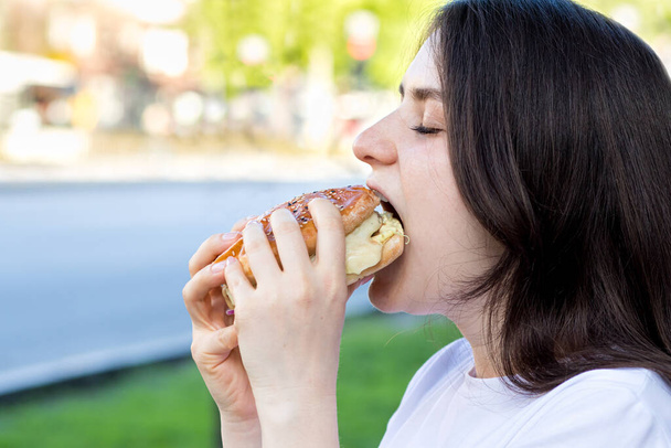 Brunette femme manger un hamburger dans la rue près de la route dans la ville. Restauration rapide sur le chemin du travail ou de l'école. Place pour le texte. - Photo, image