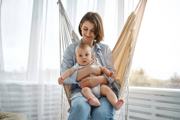 Loving mom carying of her newborn baby at home.Mother and child on a mesh hammock. Mom and baby boy playing in sunny bedroom. Parent and little kid relaxing at home. Family having fun together. - Photo, Image