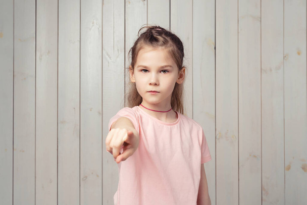 Hei, sinä. Vakava pieni tyttö 9-11-vuotias rento vaaleanpunainen t-paita osoittaa sormella kameraa, sanoo, että valitsen sinut, seisten puisen taustan yläpuolella - Valokuva, kuva