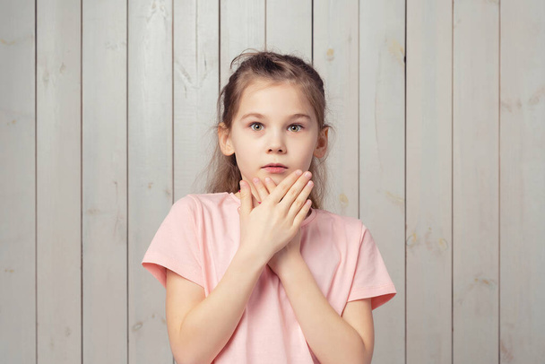 Испуганная маленькая девочка 9-11 лет в непринужденной розовой футболке, прикрывает рот ладонями и смотрит испуганно и тревожно на камеру, чего-то боится, стоя на деревянном фоне - Фото, изображение
