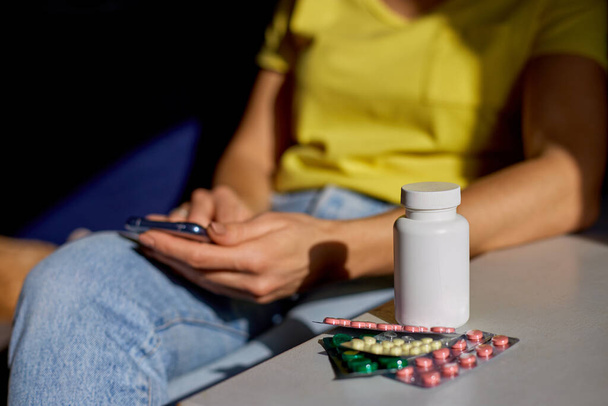 Tuntematon nainen istuu sohvalla läpipainopakkaus pillereitä online apteekki myymälä, ostaa apteekki internetissä, tilaamalla lääkkeitä verkossa älypuhelimen kotona aurinkoisena päivänä. Kova valo. Kopioi tila. - Valokuva, kuva