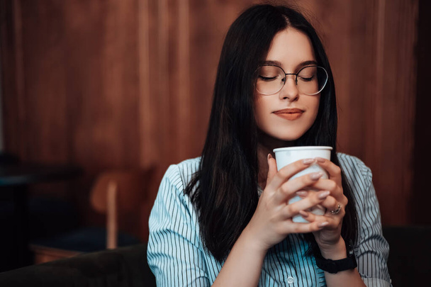 Caucasian woman sitting cafe drink plastic white cup hot beverage coffee break hold smell aroma sniffs breathe notes of caffeine. Lady dressed striped shirt stylish glasses copyspace - Photo, Image