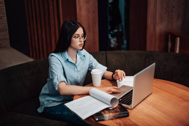 Business nainen istuu kahvila kahvi tauko avoin tila työ kannettava tietokone on lounas asiakirjoja. Lady pukeutunut raidallinen paita tyylikäs lasit puhuminen kollegoiden kanssa video ääni chat copyspace - Valokuva, kuva
