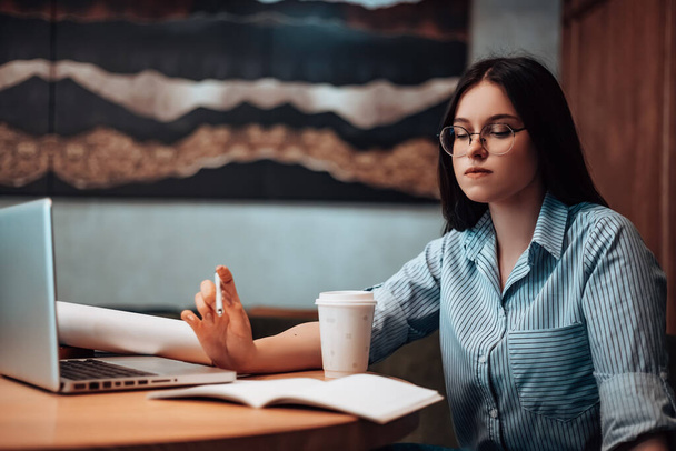 Kafede oturan küçük işletme sahibi kadın kahve içiyor açık uzay bilgisayarında öğle yemeği evrakları var. Çizgili gömlekli bayan gözlük takıyor. Şirketin gelecekteki mekânını düşünüyor. - Fotoğraf, Görsel