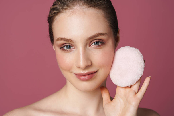 Beauty portrait of an attractive smiling sensual young woman standing isolated over pink background, using powder puff - Valokuva, kuva
