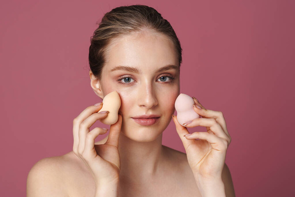 Beautiful young woman applying makeup using beauty blender sponge isolated over pink background - Foto, Imagem
