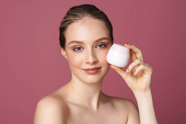 Close up beauty portrait of an attractive young woman showing jar of face cream isolated over pink background - Фото, зображення
