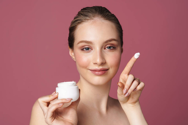 Close up beauty portrait of an attractive young woman showing jar of face cream isolated over pink background - Фото, изображение