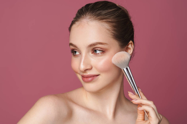 Retrato de belleza de una atractiva joven saludable sonriente que aplica maquillaje con cepillo aislado sobre fondo rosa - Foto, imagen