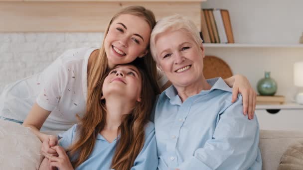 Peaceful little cute small girl sitting on sofa between loving mom and middle-aged granny, young daughter embracing mother-in-law Happy 3 three generations female family enjoying free weekend time - Footage, Video