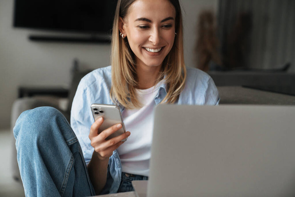 Mujer joven feliz usando el teléfono móvil mientras está sentado en casa con el ordenador portátil - Foto, Imagen