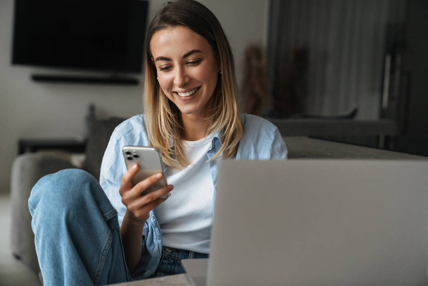 Happy young woman using mobile phone while sitting at home with laptop computer - Foto, Bild