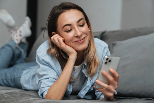 Smiling young woman using mobile phone while relaxing on a couch at home - Fotografie, Obrázek