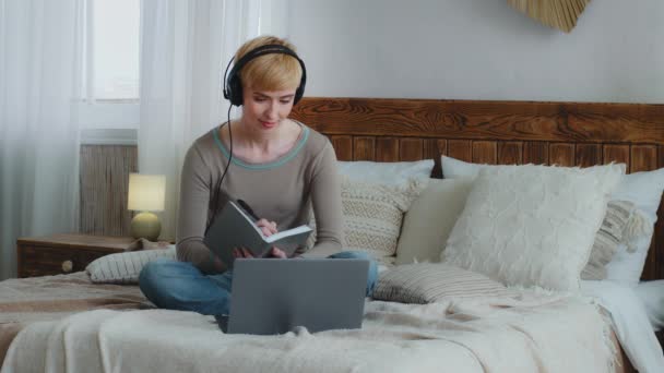 Uśmiechnięta piękna kobieta w słuchawkach zostaje w domu podczas kwarantanny patrząc na ekran laptopa, korzystając z aplikacji wideo call oglądając wykłady edukacyjne, pisząc notatki w copybooku, siedząc na wygodnym łóżku - Materiał filmowy, wideo