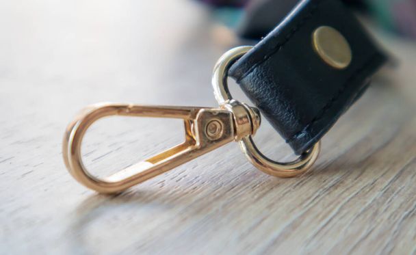 Swivel carabiner fastener with leather black bag strap on a wooden background. Metal carabiner with swivel clip or hook. Small gold fittings close-up in selective focus - Photo, Image