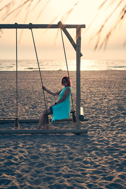 Mujer atractiva joven con el pelo rosa está descansando en balancín en la playa al atardecer. - Foto, Imagen