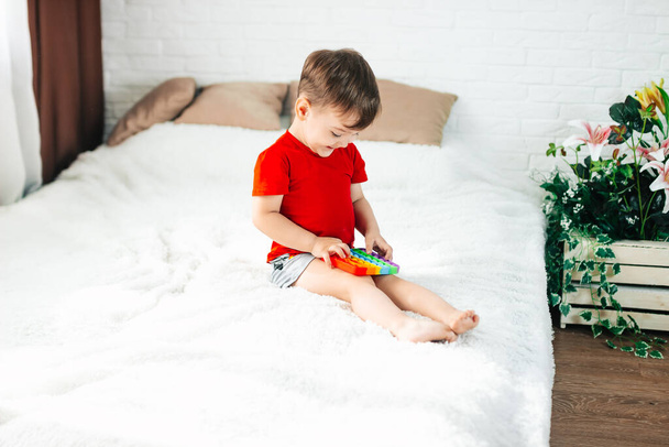 Kleine baby jongen zit thuis op het bed, spelen Pop-it nieuwe anti-stress speelgoed, populair bij kinderen, helpt hen om zich te concentreren. Baby spelen met een pop-it anti-stress speelgoed. - Foto, afbeelding
