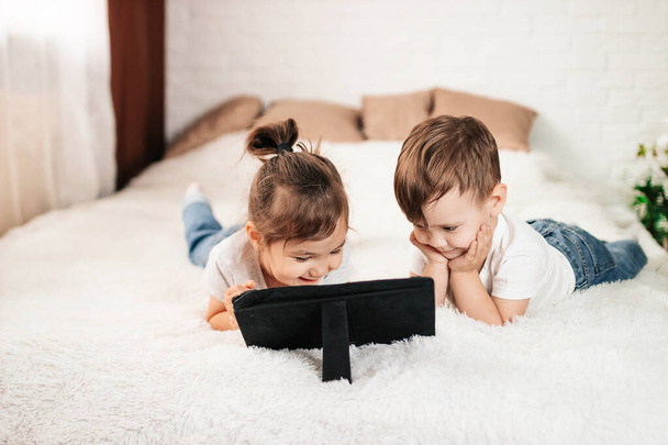 Δύο παιδιά χρησιμοποιούν έναν υπολογιστή ταμπλέτας σε ένα λευκό κρεβάτι. Ένα αγόρι και ένα κορίτσι με ένα tablet υπολογιστή σε ένα λευκό δωμάτιο - Φωτογραφία, εικόνα