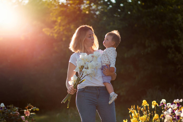 Portret van een blanke moeder met een boeket irissen en haar kleine meisje in haar armen. Zonsondergang licht in de tuin. Het concept van 8 maart en Happy Children 's Day. - Foto, afbeelding