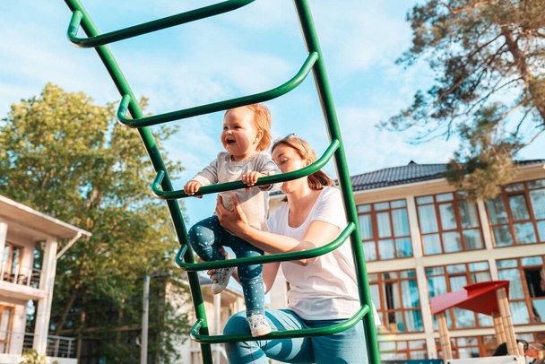 Uma jovem mãe caucasiana brinca com uma criança no parque infantil, ensina o bebê a subir as escadas. Férias de verão e de família. - Foto, Imagem