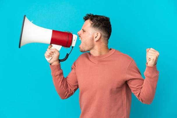Young caucasian handsome man isolated on blue background shouting through a megaphone to announce something in lateral position - Fotografie, Obrázek