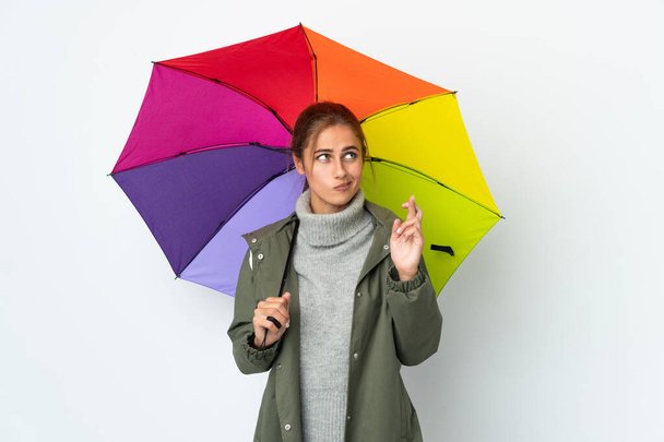 Young woman holding an umbrella isolated on white background with fingers crossing and wishing the best - Photo, Image