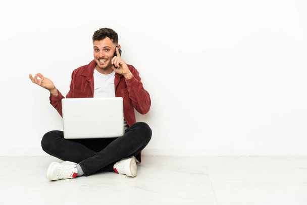 Genç, yakışıklı, beyaz bir adam dizüstü bilgisayarla yerde oturuyor ve cep telefonuyla sohbet ediyor. - Fotoğraf, Görsel