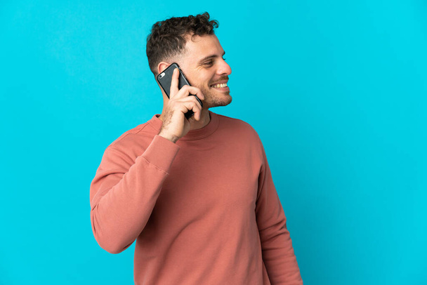 Young caucasian handsome man isolated on blue background keeping a conversation with the mobile phone with someone - Foto, immagini