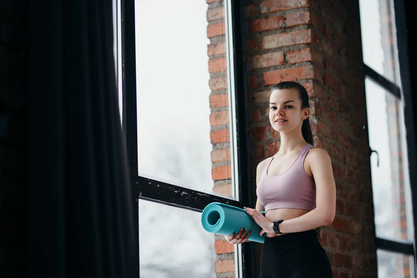 Cute young girl fitness trainer holding yoga mat in hand while standing in the gym and getting ready for workout - Zdjęcie, obraz