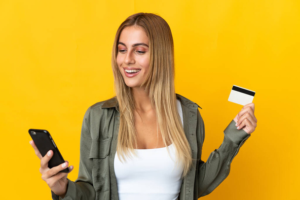 Young blonde woman isolated on yellow background buying with the mobile with a credit card - Photo, Image