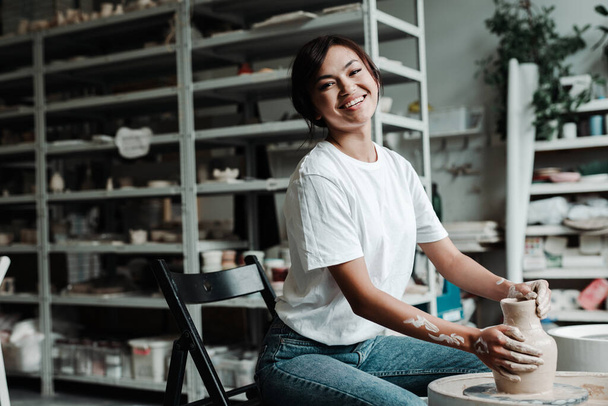 Beautiful happy asian girl making a clay vase in a workshop smiling and looking at the camera - Foto, imagen