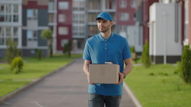 Modrý doručovatel pošle zákazníkovi balíček, než doručí náklad. 4k rozlišení a zpomalený záběr. Muž pošťák s balíkem v rukou na ulici. Poštovní koncept. Doručení služby. - Záběry, video