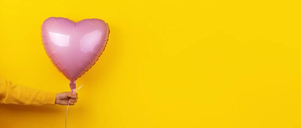 różowy balon foliowy w kształcie serca w dłoni nad żółtym tłem, układ panoramiczny - Zdjęcie, obraz