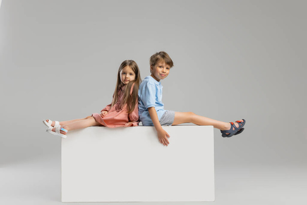 İki çocuk, ağabeyi ve kız kardeşi gri stüdyo arka planında izole bir şekilde kutuda oturuyorlar.. - Fotoğraf, Görsel