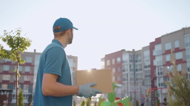 Een bezorger in een blauw T-shirt en pet draagt een kartonnen doos in de stad op zoek naar het adres van de bezorger. Leverancier gaat met een doos in slow motion - Video