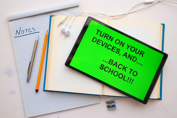 Tablica z napisem "włącz swoje urządzenia... i wracaj do szkoły", na ekranie, w czerni na zielonym tle, a obok notatniki, długopis, ołówek, temperówka i słuchawki. Początki szkół i e-learning. - Zdjęcie, obraz
